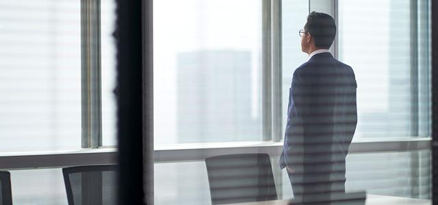 Business professional in a suit standing by office window, looking at city skyline in a modern workspace.